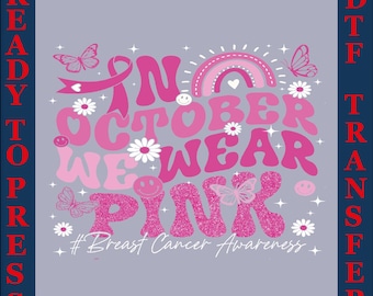 Op de afbeelding: Een kant-en-klare DTF-transfer met een grijze achtergrond en een roze ontwerp. Het ontwerp bevat de tekst "In October We Wear Pink" met een lint, regenboog, vlinders en madeliefjes. De zin "Breast Cancer Awareness" is ook opgenomen.