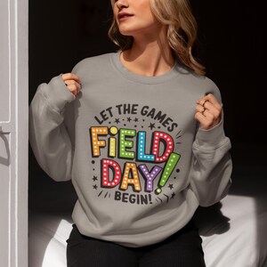 May include: A gray sweatshirt with the text "Let the games begin!" and a colorful graphic that says "Field Day!" in a retro-style font.