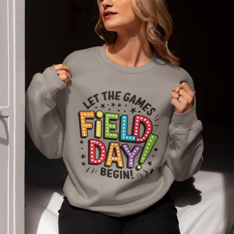 May include: A gray sweatshirt with the text "Let the games begin!" and a colorful graphic that says "Field Day!" in a retro-style font.