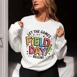 May include: White sweatshirt with a colorful graphic that says "Let the games begin! Field Day!" The graphic is in a retro style with light bulbs around the letters.