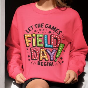 May include: Pink sweatshirt with the text "Let the games begin!" and "Field Day!" in colorful, outlined letters with light bulbs around the words.