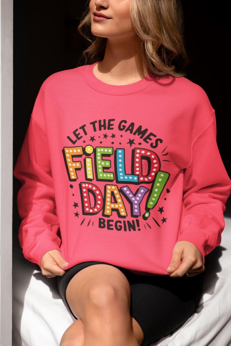 May include: Pink sweatshirt with the text "Let the games begin!" and "Field Day!" in colorful, outlined letters with light bulbs around the words.
