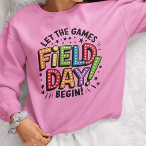 May include: Pink sweatshirt with the text "Let the games begin!" and "Field Day!" in colorful, outlined letters with light bulbs around the words.