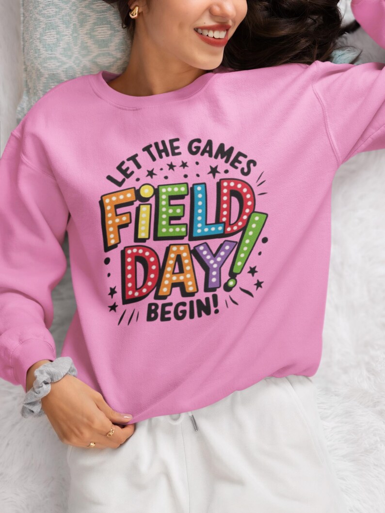 May include: Pink sweatshirt with the text "Let the games begin!" and "Field Day!" in colorful, outlined letters with light bulbs around the words.