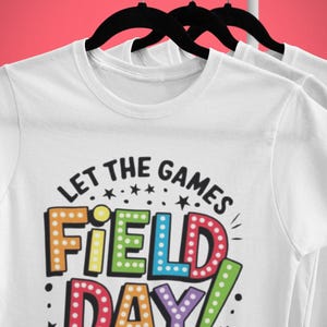 May include: White t-shirt with a colorful graphic that says "Let the games begin! Field Day!" The graphic is in a retro style with light bulbs around the letters.