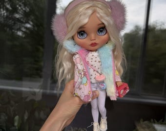 Può includere: Una bambola con lunghi capelli biondi indossa paraorecchie rosa soffici, una giacca rosa con motivo a nuvole e stelle, un top bianco con stelle, leggings bianchi e stivaletti color crema. La bambola porta un piccolo accessorio rosa e una bottiglia.