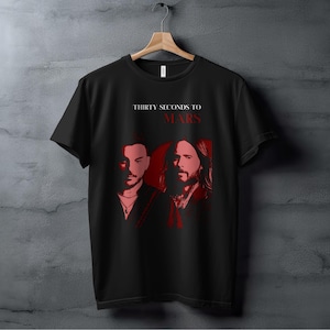 Puede incluir: Camiseta negra con un gráfico rojo de la banda Thirty Seconds to Mars. El nombre de la banda está impreso en blanco sobre el gráfico.