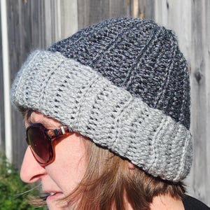 Op de afbeelding: Een handgemaakte, tweekleurige beanie-muts. Het bovenste gedeelte is donkergrijs, terwijl de omgeslagen rand lichtgrijs is. De hoed is gemaakt van een gebreid materiaal en is ontworpen om het hoofd warm te houden.