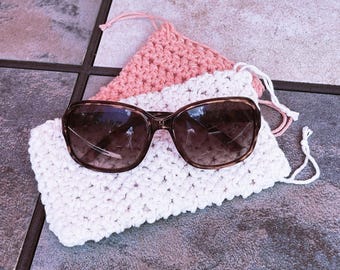 May include: Sunglasses with brown frames rest on a white crocheted pouch, with a pink crocheted pouch behind it. The sunglasses have a gradient tint. The pouches have drawstrings. The items are on a tiled surface.