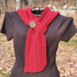 Puede incluir: Una bufanda de crochet roja con un botón de madera. La bufanda está atada alrededor de una camisa negra.