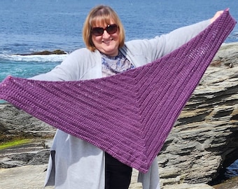 May include: A purple crocheted shawl with a triangular shape, held by a person standing on a rocky shore.