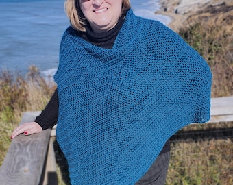 Op de afbeelding: Een tealblauwe gebreide poncho met een v-hals. De poncho wordt gedragen door een persoon die op een houten leuning staat met uitzicht op een waterpartij.