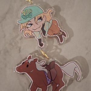 Puede incluir: Un llavero de acrílico de dos capas con un cierre dorado en forma de estrella. El dije superior muestra un personaje rubio con una gorra verde azulado con el texto DIO, y el dije inferior muestra un caballo marrón con silla de montar.