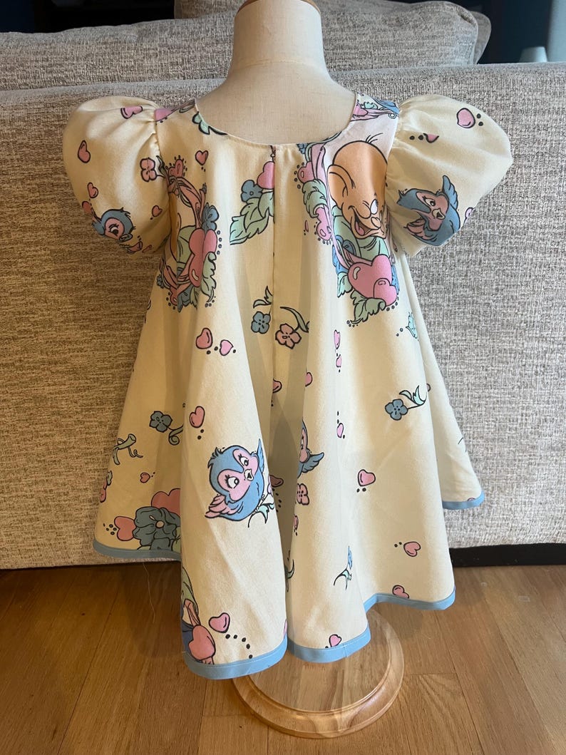 May include: A cream-coloured dress with a blue trim. The dress has a cartoon print of birds and hearts.