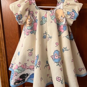 May include: A light yellow dress with a blue trim. The dress features a print of cartoon characters, including birds, hearts, and flowers.