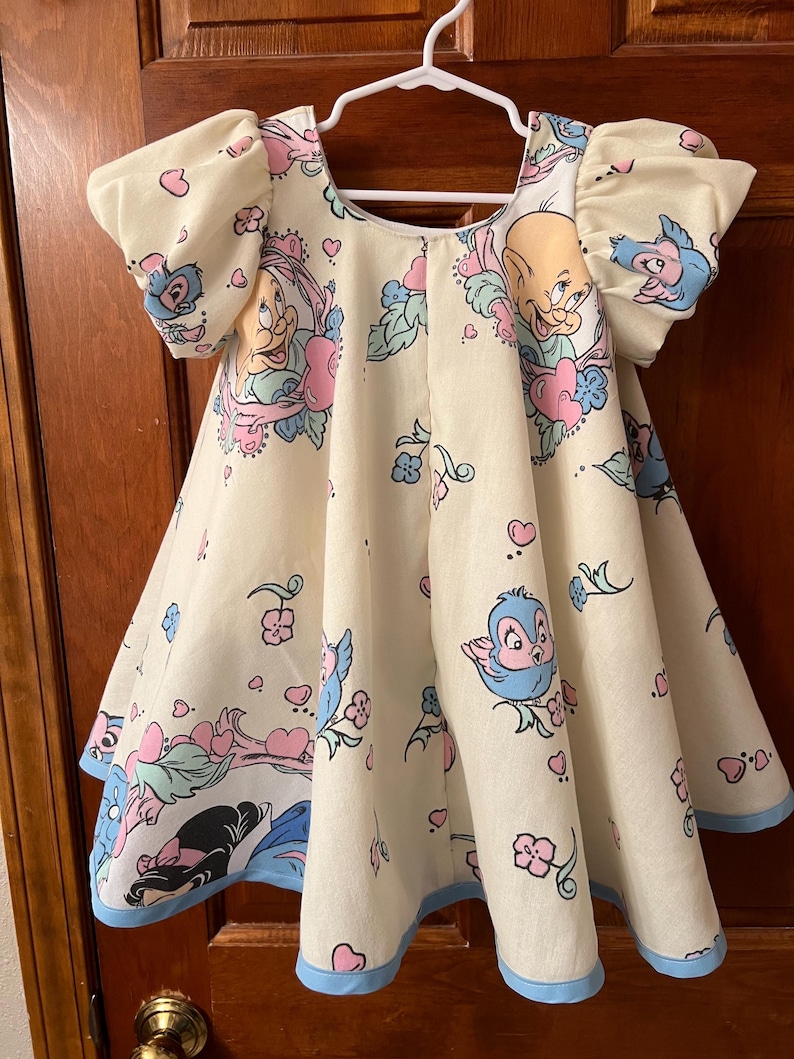 May include: A light yellow dress with a blue trim. The dress features a print of cartoon characters, including birds, hearts, and flowers.