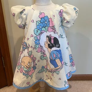 May include: A white dress with a blue and pink floral print featuring Snow White and Dopey. The dress has puffed sleeves and a blue trim around the bottom.