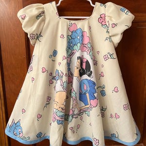 May include: A white dress with a blue trim and a print of Snow White and the Seven Dwarfs. The dress has puffed sleeves and a scoop neck.