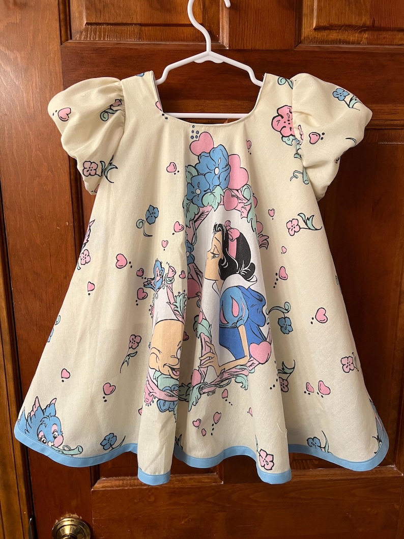 May include: A white dress with a blue trim and a print of Snow White and the Seven Dwarfs. The dress has puffed sleeves and a scoop neck.