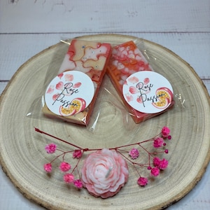 Peut inclure: Deux petites fondantes de cire rose et blanche avec un motif floral. Les fondantes de cire sont emballées dans des sachets en plastique transparent avec une étiquette ronde qui dit "Rose Passion". Une petite bougie en forme de fleur rose est au centre de l'image.
