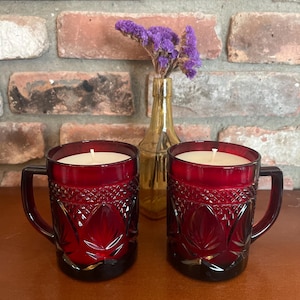 May include: Two ruby red glass mugs filled with white wax candles. The mugs have a decorative pattern and handles. A small gold vase with purple flowers sits behind the mugs, with a brick wall in the background.