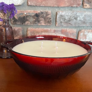 May include: A large, round, red glass bowl candle with three wicks. The candle has a creamy white wax and is set in a decorative bowl with handles. A small purple glass bottle with purple flowers is to the left.