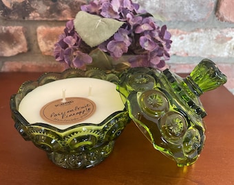 May include: A green glass candle holder with a lid, containing a white candle with three wicks. The candle has a brown label that reads "Passionfruit Pineapple Handmade with". Purple flowers are in the background.