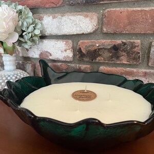 May include: A large, leaf-shaped, dark green candle holder filled with a cream-colored candle. The candle has multiple wicks and a small, round label. A small vase with white and green flowers sits to the left of the candle holder.