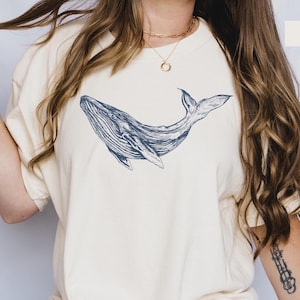 May include: A woman wearing a cream colored t-shirt with a blue whale graphic. The shirt is oversized and has a relaxed fit. The woman is wearing blue jeans and has long brown hair.