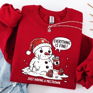 May include: Red crewneck sweatshirt with a snowman graphic. The snowman is melting, wearing a Santa hat, and holding a coffee cup. Text reads "Everything is Fine!" and "Just Having a Meltdown."