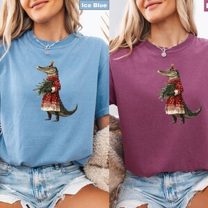 Pu&ograve; includere: T-shirt blu ghiaccio e color bacca con un'illustrazione stravagante di un alligatore vestito con un cappotto rosso con finiture bianche e un cappello festivo, che tiene un piccolo albero di Natale. L'alligatore &egrave; l'elemento centrale del design.