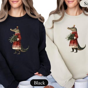 Pu&ograve; includere: Felpe nere e color sabbia con un'illustrazione stravagante di un alligatore vestito con un abito rosso festivo, che tiene in mano un fascio di rami sempreverdi. L'alligatore indossa un cappello a stella, aggiungendo un tocco festoso al design.