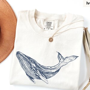 May include: A cream colored t-shirt with a blue whale graphic and the text "COMFORT COLORS" on the tag. The shirt is laid flat on a white surface with a brown hat and a dried palm leaf.