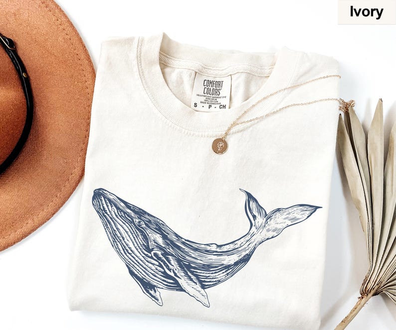 May include: A cream colored t-shirt with a blue whale graphic and the text "COMFORT COLORS" on the tag. The shirt is laid flat on a white surface with a brown hat and a dried palm leaf.