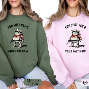 Puede incluir: Dos sudaderas, una verde militar y otra rosa claro, cada una con una rana de dibujos animados con sombrero de vaquero y sosteniendo una pistola. El texto "YOU JUST YEE'D YOUR LAST HAW" est&aacute; encima y debajo de la ilustraci&oacute;n de la rana.