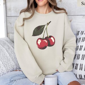 K&ouml;nnte beinhalten: Sandfarbenes Sweatshirt mit einer Grafik von zwei reifen roten Kirschen und einem gr&uuml;nen Blatt. Die Kirschen haben ein gl&auml;nzendes Finish. Das Sweatshirt ist ein Rundhals-Stil und besteht aus einem weichen Material.