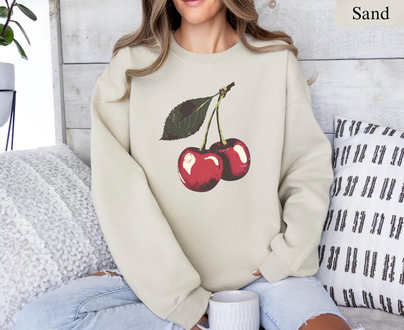 K&ouml;nnte beinhalten: Sandfarbenes Sweatshirt mit einer Grafik von zwei reifen roten Kirschen und einem gr&uuml;nen Blatt. Die Kirschen haben ein gl&auml;nzendes Finish. Das Sweatshirt ist ein Rundhals-Stil und besteht aus einem weichen Material.
