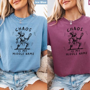May include: Ice blue and berry-colored t-shirts with the text "CHAOS IS MY MIDDLE NAME." Each shirt features a black illustration of a frog chef holding a whisk. The shirts are short-sleeved and have a relaxed fit.