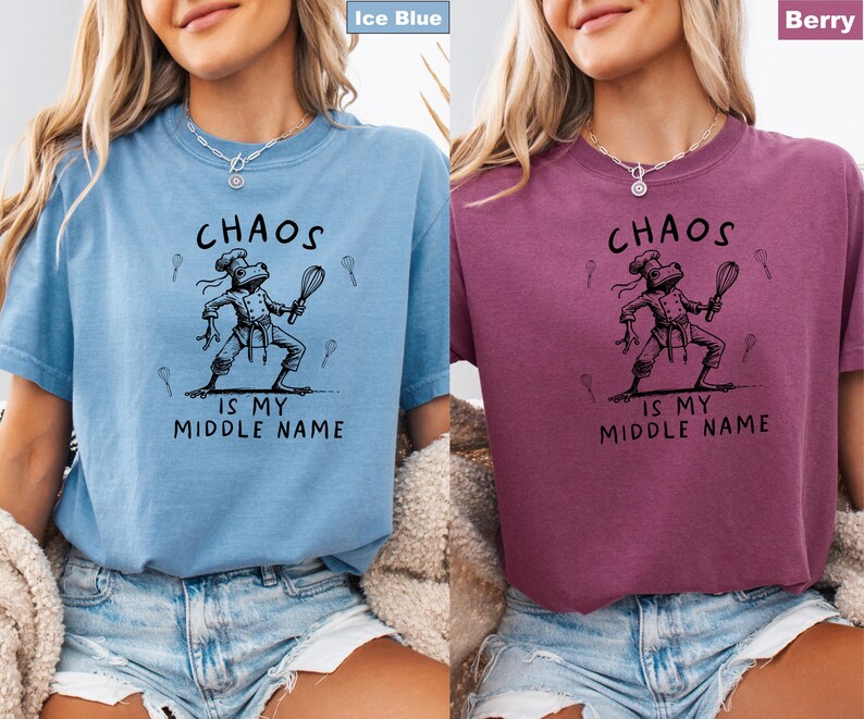 May include: Ice blue and berry-colored t-shirts with the text "CHAOS IS MY MIDDLE NAME." Each shirt features a black illustration of a frog chef holding a whisk. The shirts are short-sleeved and have a relaxed fit.