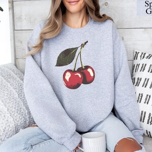 K&ouml;nnte beinhalten: Hellgraues Sport Grey Sweatshirt mit einer Grafik von zwei roten Kirschen und gr&uuml;nen Bl&auml;ttern. Der Text "Sport Grey" befindet sich oben rechts. Ein l&auml;ssiger Stil.