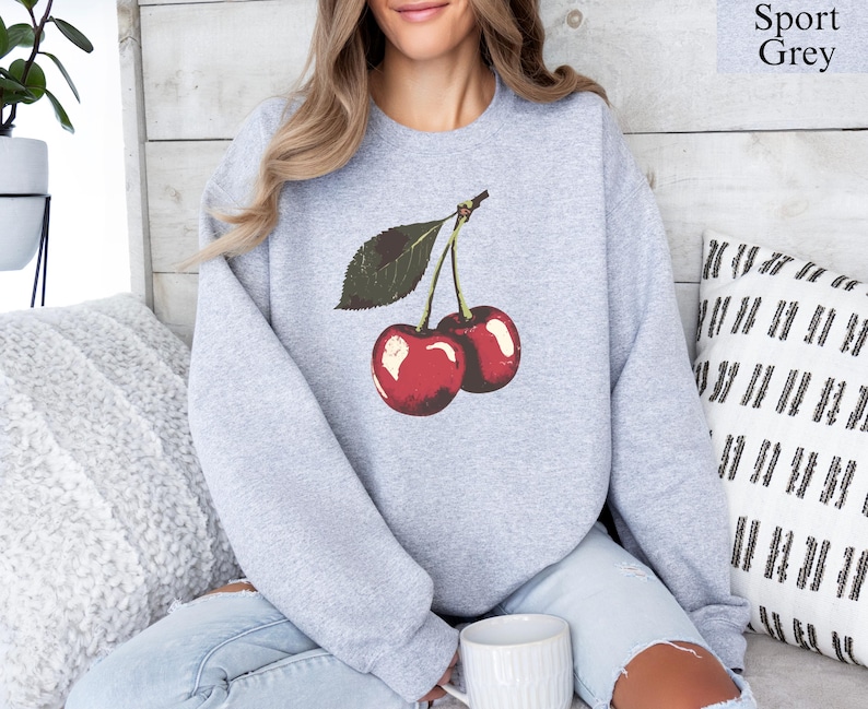 K&ouml;nnte beinhalten: Hellgraues Sport Grey Sweatshirt mit einer Grafik von zwei roten Kirschen und gr&uuml;nen Bl&auml;ttern. Der Text "Sport Grey" befindet sich oben rechts. Ein l&auml;ssiger Stil.