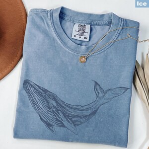 May include: A light blue t-shirt with a whale graphic printed on the front. The shirt has a distressed look and is made of a soft, comfortable fabric. The text "COMFORT COLORS" is printed on the tag.