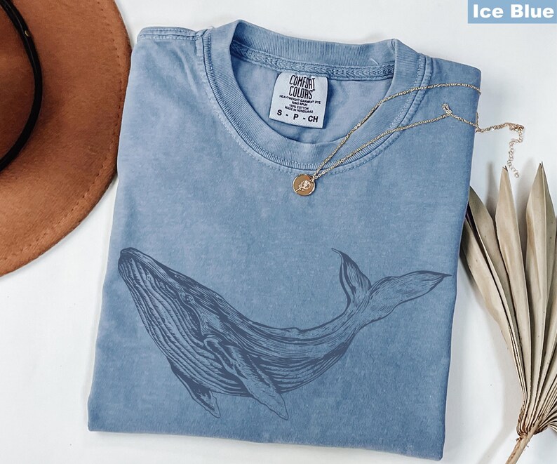 May include: A light blue t-shirt with a whale graphic printed on the front. The shirt has a distressed look and is made of a soft, comfortable fabric. The text "COMFORT COLORS" is printed on the tag.