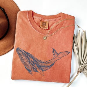 May include: A coral-colored t-shirt with a blue whale graphic. The shirt is made of a soft, comfortable fabric and has a relaxed fit.