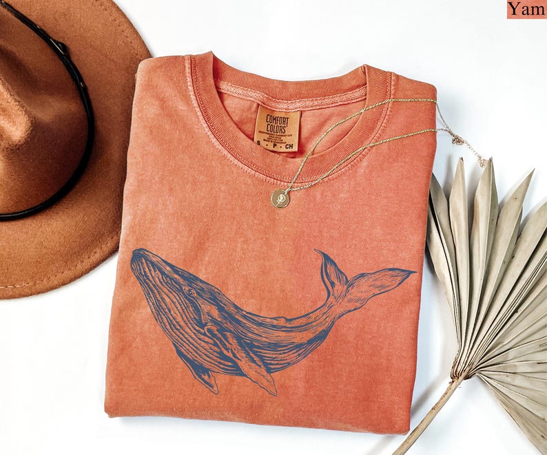 May include: A coral-colored t-shirt with a blue whale graphic. The shirt is made of a soft, comfortable fabric and has a relaxed fit.