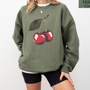 K&ouml;nnte beinhalten: Milit&auml;rgr&uuml;nes Sweatshirt mit einer Grafik von zwei roten Kirschen und einem gr&uuml;nen Blatt. Das Sweatshirt hat einen Rundhalsausschnitt und lange &Auml;rmel. Der Text "Military Green" befindet sich in der oberen rechten Ecke.