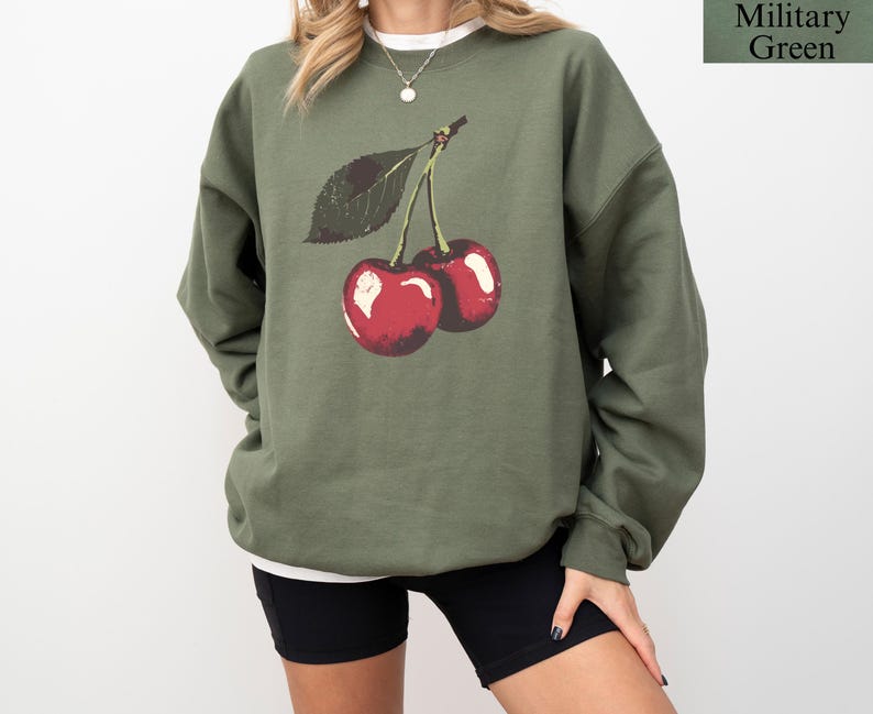 K&ouml;nnte beinhalten: Milit&auml;rgr&uuml;nes Sweatshirt mit einer Grafik von zwei roten Kirschen und einem gr&uuml;nen Blatt. Das Sweatshirt hat einen Rundhalsausschnitt und lange &Auml;rmel. Der Text "Military Green" befindet sich in der oberen rechten Ecke.