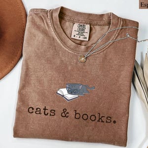 K&ouml;nnte beinhalten: Espressofarbenes T-Shirt mit Rundhalsausschnitt und kurzen &Auml;rmeln. Das Shirt zeigt eine Grafik einer grauen Katze, die auf einem offenen Buch liegt, mit dem Schriftzug "cats & books." darunter. Eine silberne Halskette mit kleinem Anh&auml;nger liegt auf dem Shirt.