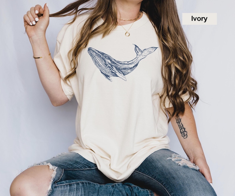 May include: A woman wearing a cream colored t-shirt with a blue whale graphic. The shirt is oversized and has a relaxed fit. The woman is wearing blue jeans and has long brown hair.