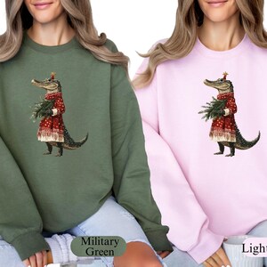 Pu&ograve; includere: Due felpe, una verde militare e una rosa chiaro, ciascuna con una stravagante illustrazione di un alligatore vestito con un abito rosso festivo, che tiene in mano un mazzo di verde. L'alligatore indossa una corona d'oro a forma di stella. Le felpe sono casual.